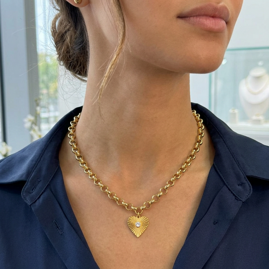 A close-up of a woman wearing a gold heart pendant necklace with a small white pearl accent, paired with a navy blue collared shirt.