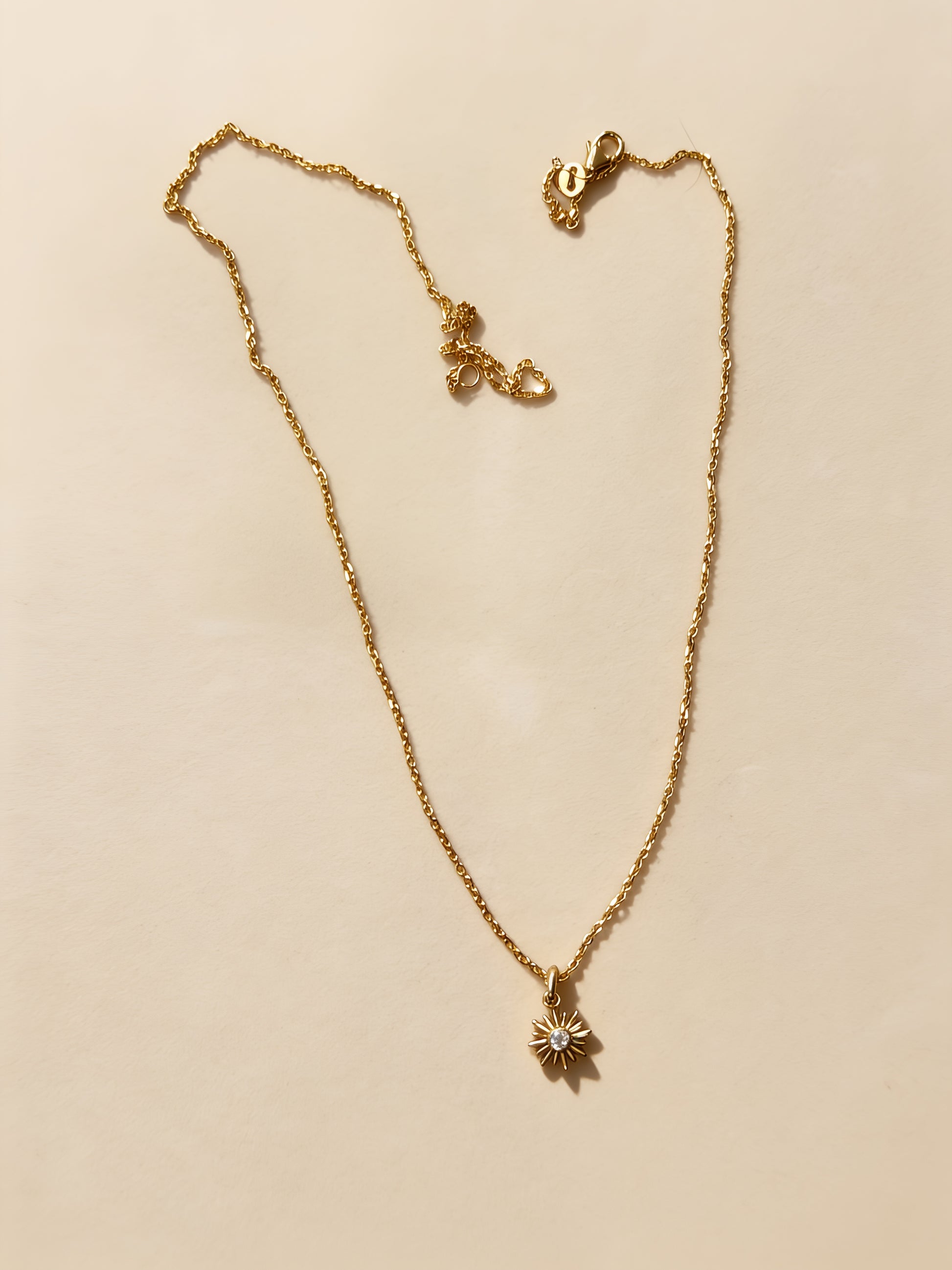 A delicate gold chain necklace with a small starburst pendant, laid flat on a warm beige background.